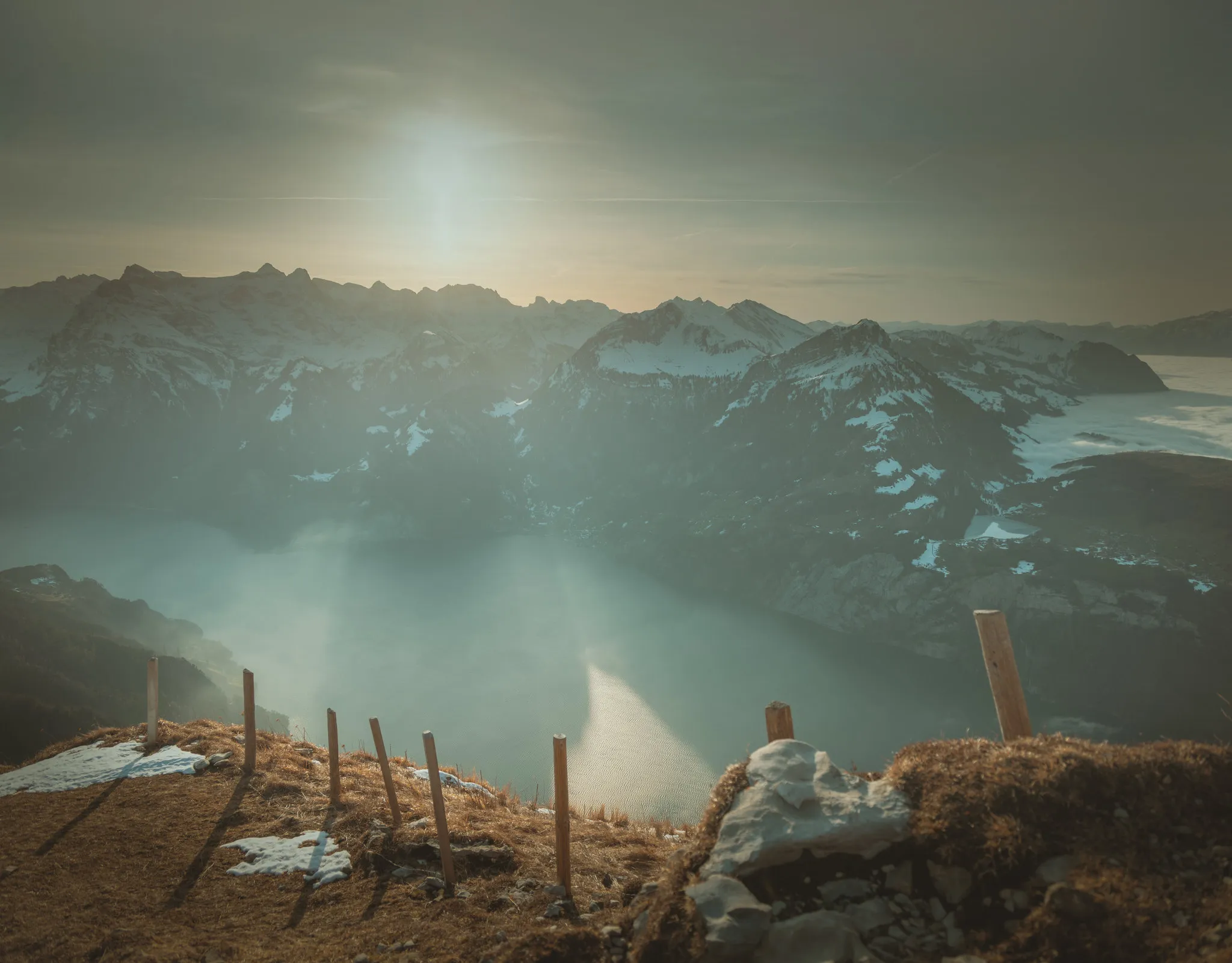 Lake Lucerne far below a mountain ridge at Stoos, with snow-covered peaks in hazy sunlight